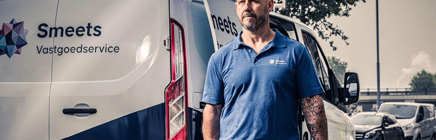 Een medewerker met een blauwe polo staat naast een bedrijfsbus op een parkeerplaats, met verkeer en bomen op de achtergrond.