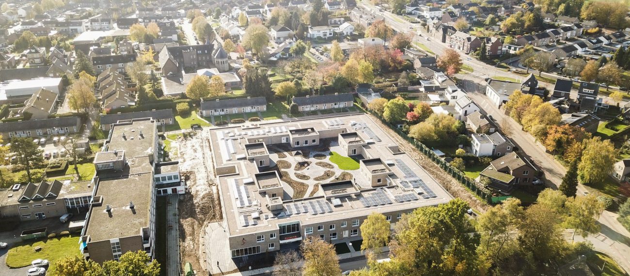 Luchtfoto van een groot gebouw met binnenplaats en zonnepanelen, omringd door woonhuizen en groen.