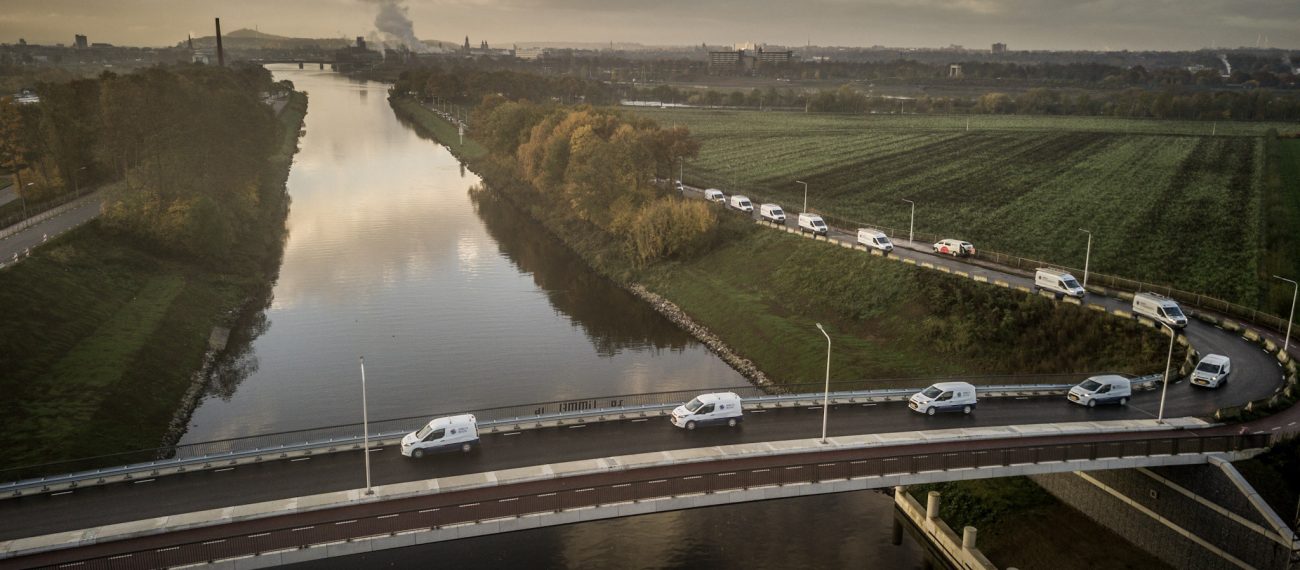 Luchtfoto van een rivier met een brug in de voorgrond en een colonne witte bedrijfsbusjes die over de kronkelende weg langs het water rijden door een landelijk landschap bij zonsopkomst.
