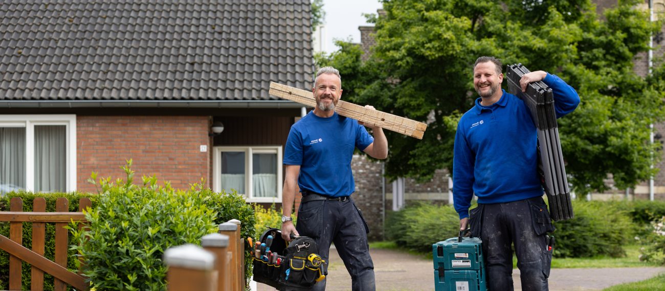 Twee vaklieden met gereedschap lopen buiten voor een woning.