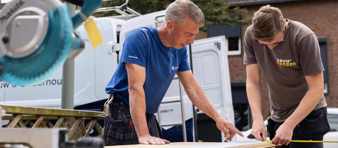 Twee medewerkers meten en markeren een houten plaat buiten; één persoon wijst op de maat terwijl de ander een meetlint vasthoudt, met een werkbus en huizen op de achtergrond.
