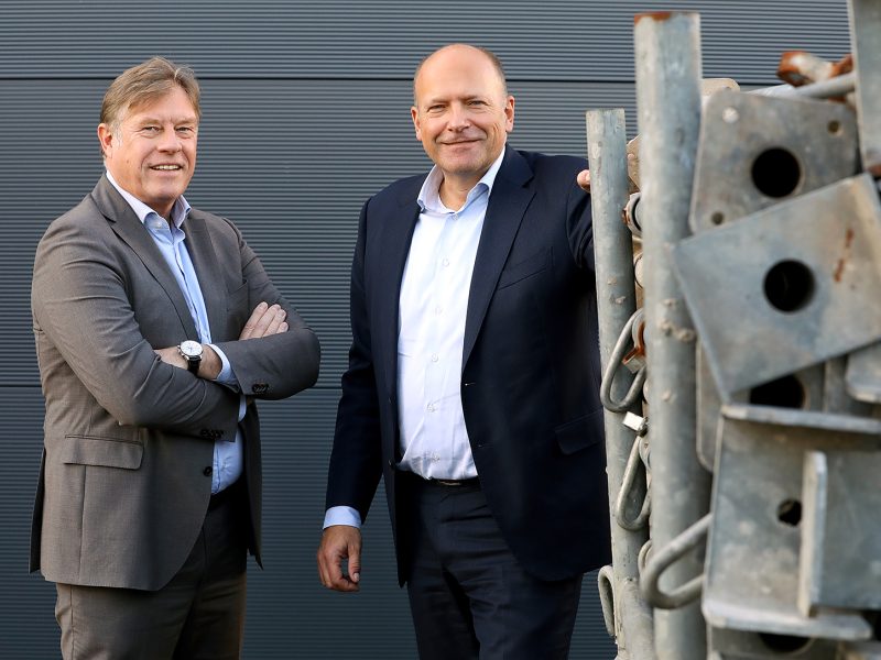 Twee mannen in pak poseren lachend buiten voor een grijze wand, naast een stapel metalen bouwmaterialen
