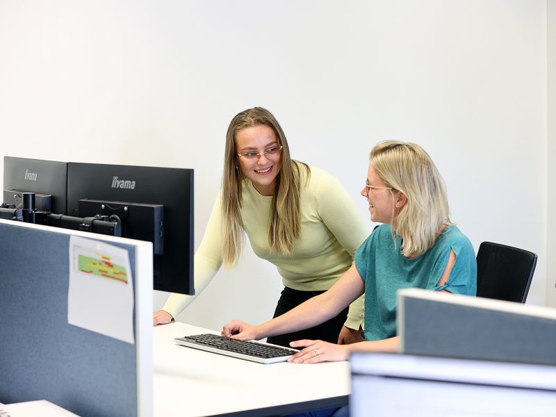 Twee collega’s achter een bureau die lachend samenwerken achter een computer in een kantooromgeving.