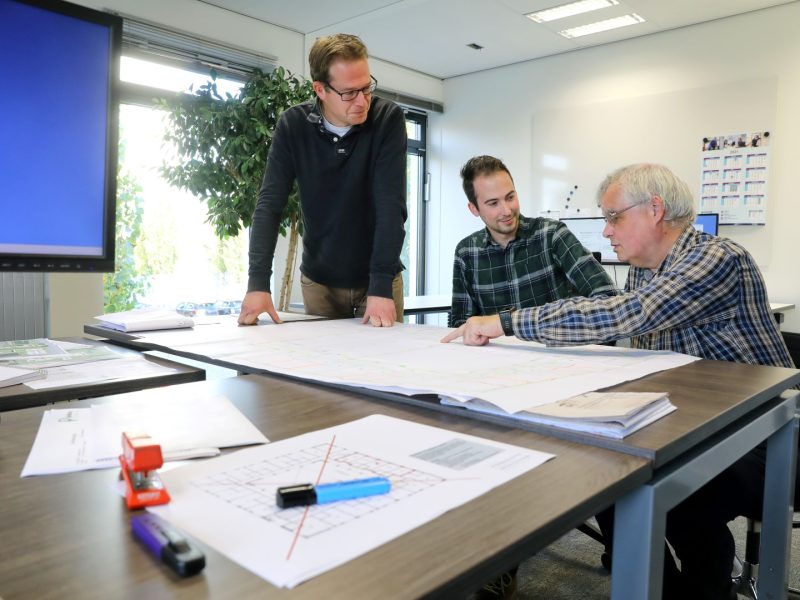 Drie mannen overleggen in een kantoorruimte boven bouwtekeningen op tafel