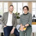 Twee mensen poseren vriendelijk voor de camera terwijl zij op een bureau naast elkaar zitten