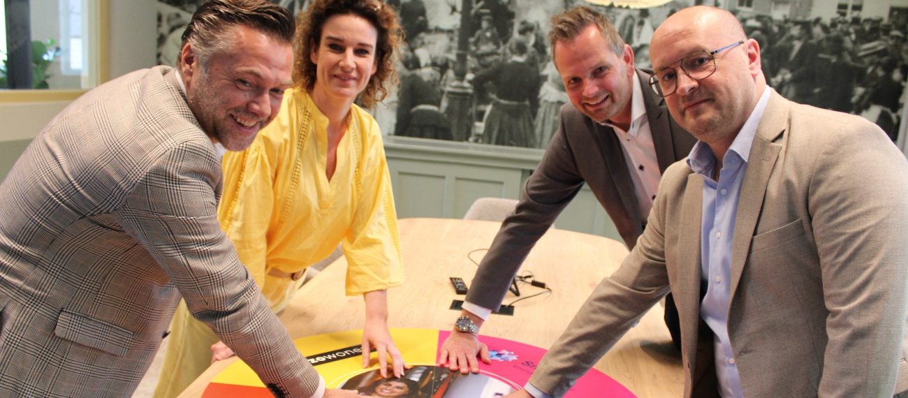 Vier mensen leggen hun handen op een kleurrijk cirkeldiagram op tafel tijdens een overleg.