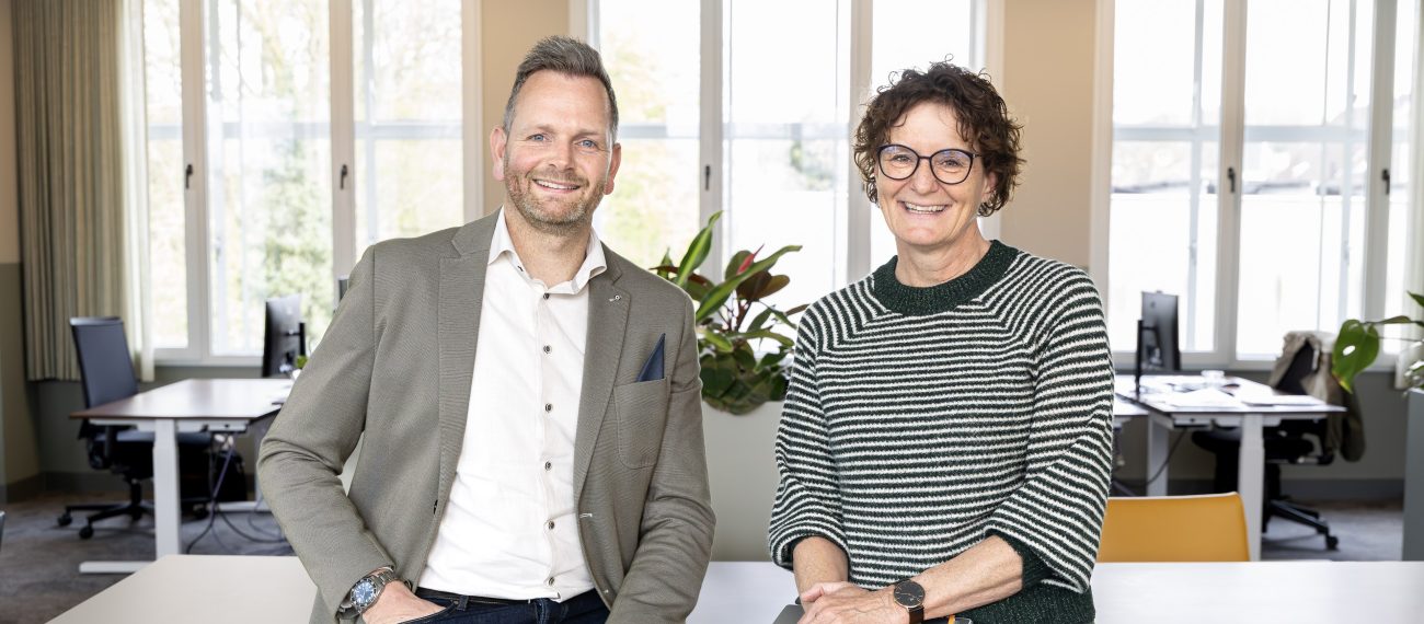 Twee mensen poseren vriendelijk voor de camera terwijl zij op een bureau naast elkaar zitten