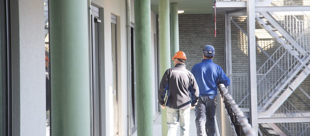 Bouwmedewerkers met helmen lopen over een galerij van een nieuwbouwcomplex langs groene kolommen.