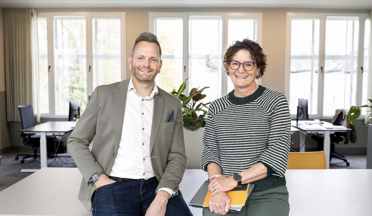 Twee mensen poseren vriendelijk voor de camera terwijl zij op een bureau naast elkaar zitten