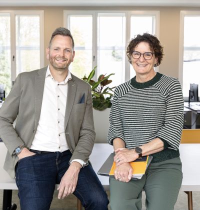Twee mensen poseren vriendelijk voor de camera terwijl zij op een bureau naast elkaar zitten