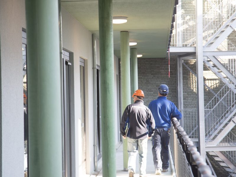 Bouwmedewerkers met helmen lopen over een galerij van een nieuwbouwcomplex langs groene kolommen.