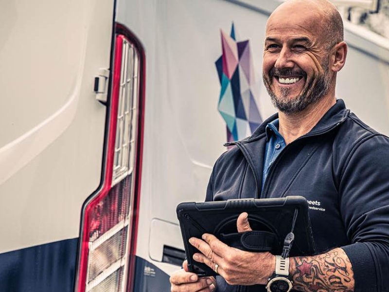 Glimlachende man bij een bedrijfsbus houdt een tablet vast tijdens zijn werk buiten.